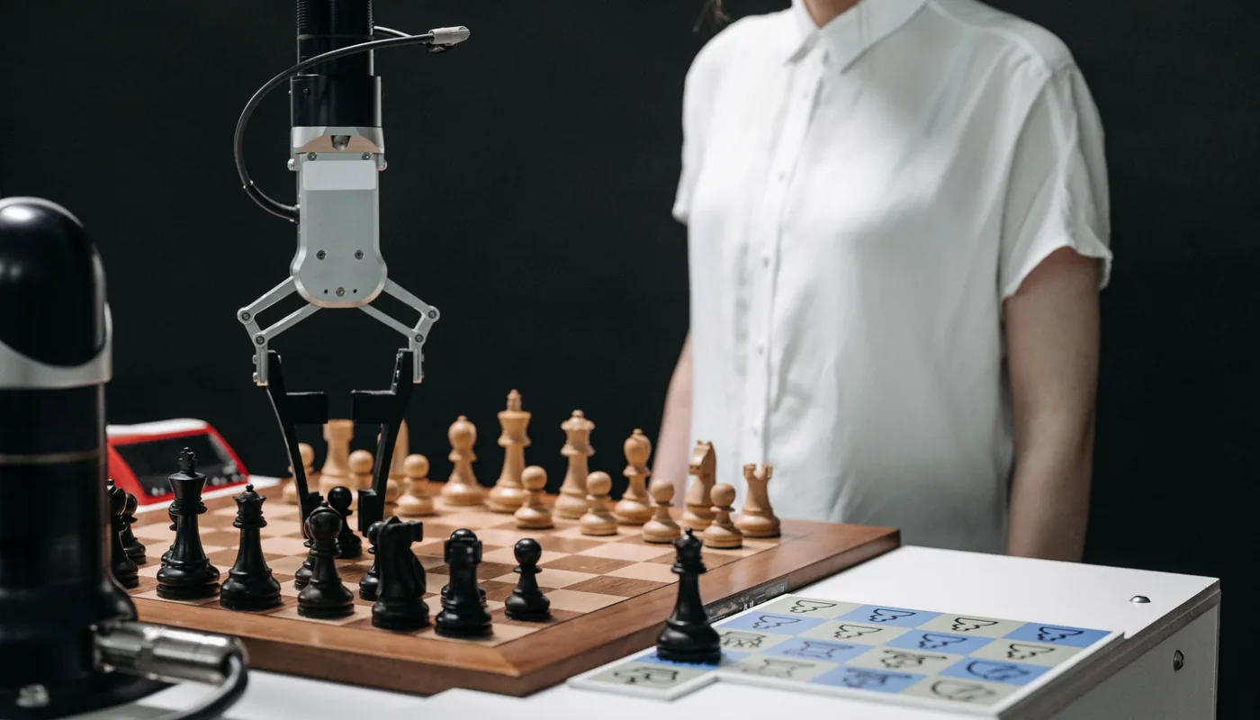 A robotic arm with a gripper hovers over a chess board mid-game while a person in a white shirt stands beside it. Automation is fast, but someone still has to decide the strategy.
